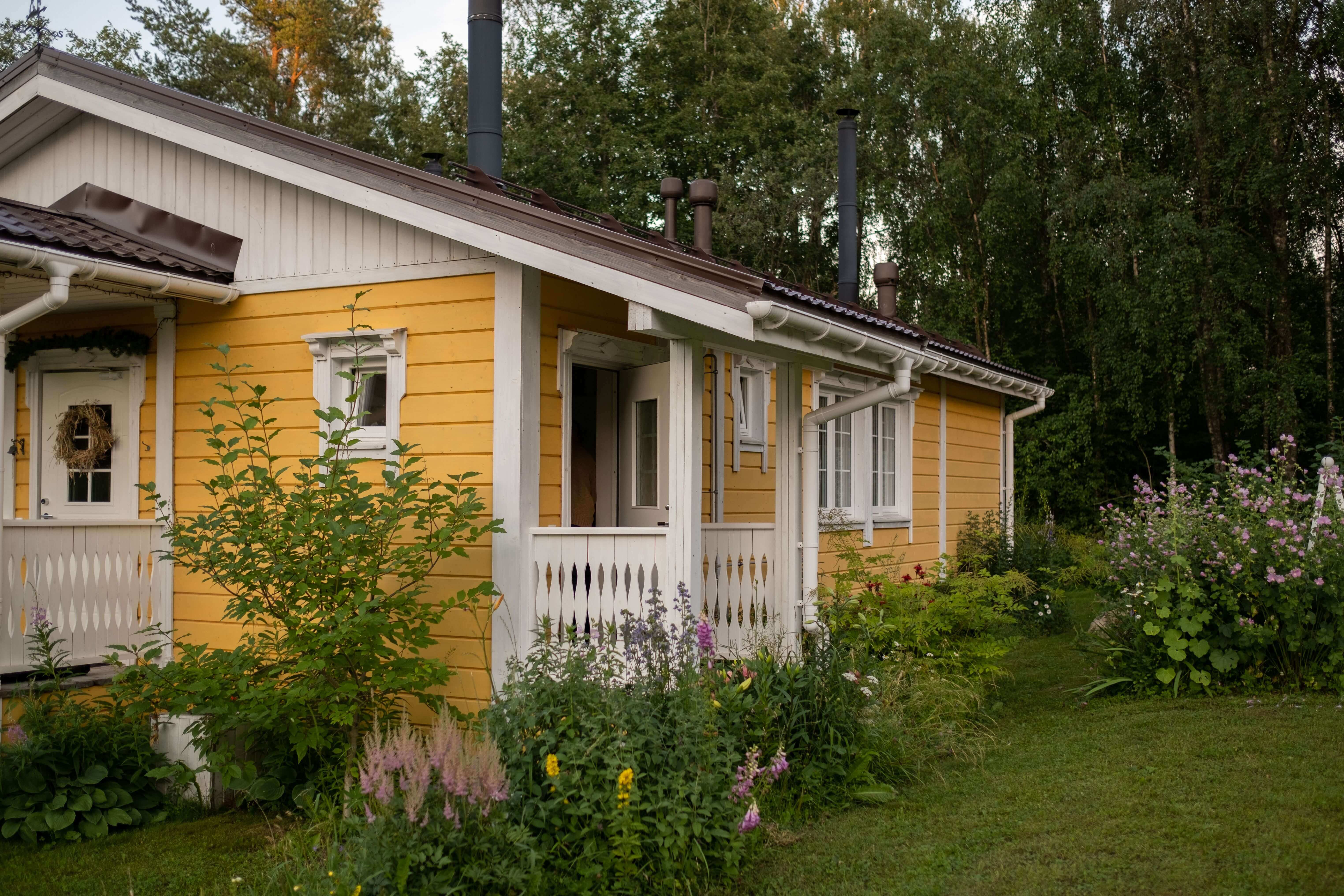 Gîte en bois jaune avec finitions blanches entouré d'un jardin fleuri et bordé par la forêt