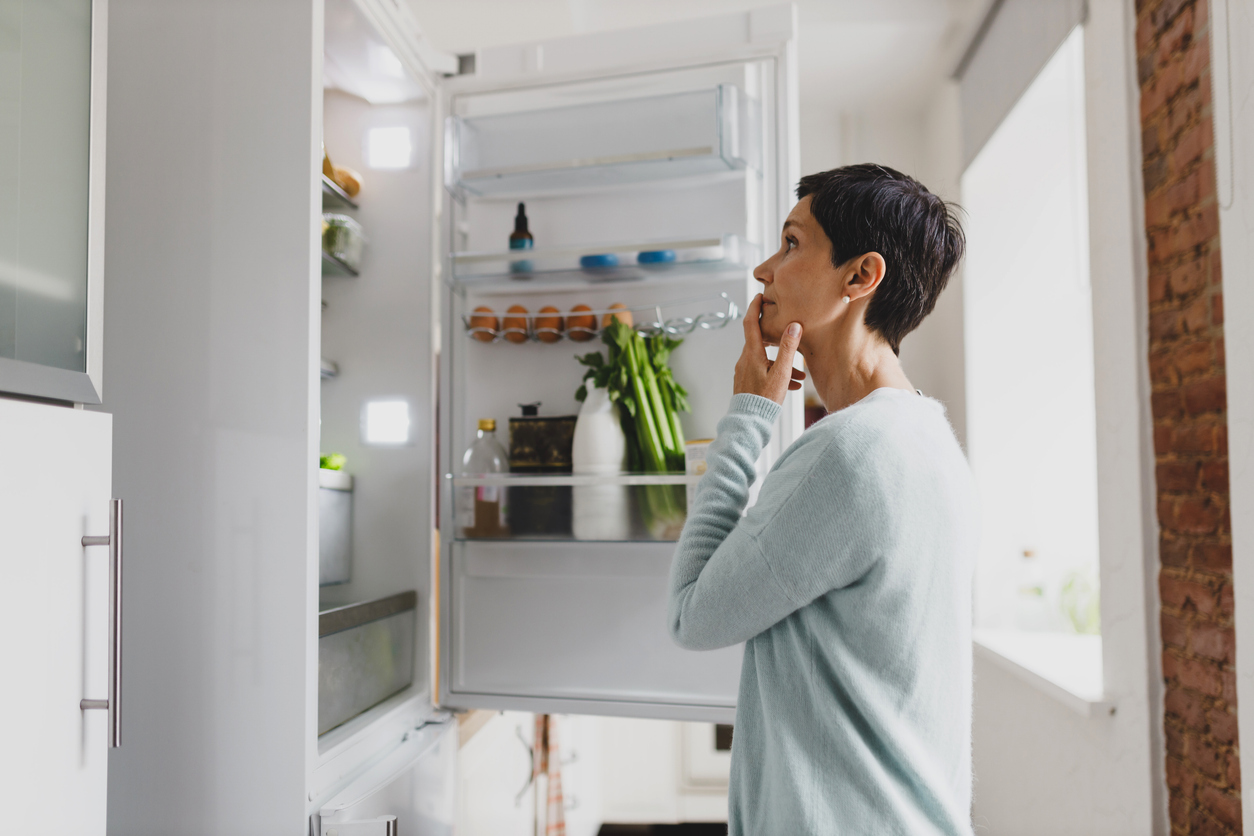 Side view of mature female with short hair standing in front of opened refrigerator at home with puzzled pensive facial expression, thinking of cooking breakfast, looking for ingredients