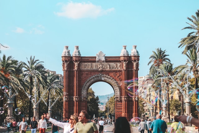 Tourists walking along a sunny boulevard in front of the Arc de Triomf in Barcelona, Spain, with palm trees and soap bubbles in the foreground.
