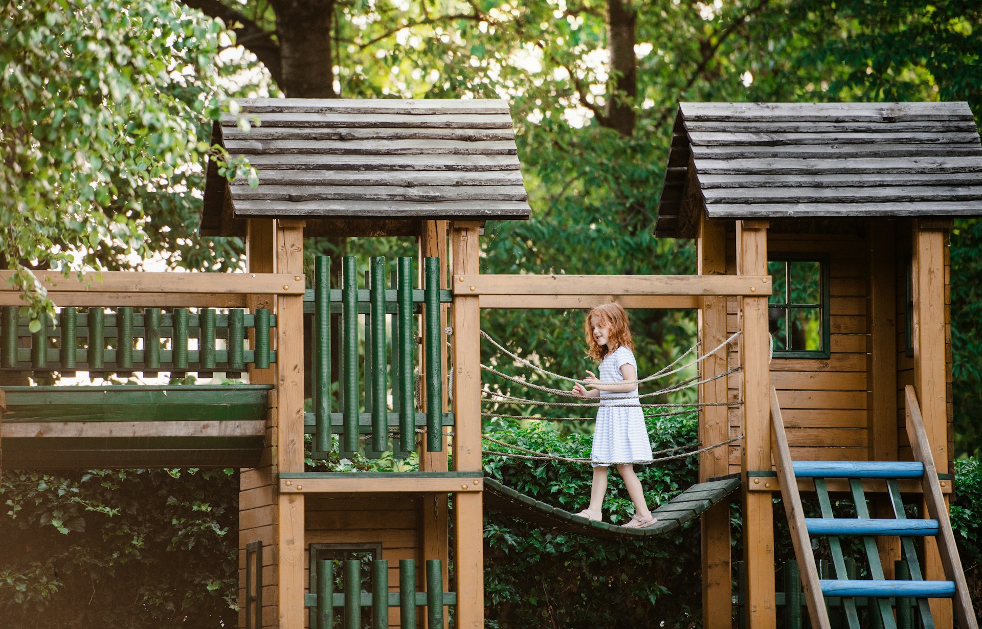 Young girl walking across a rope bridge on a wooden outdoor play structure in a holiday rental garden surrounded by trees