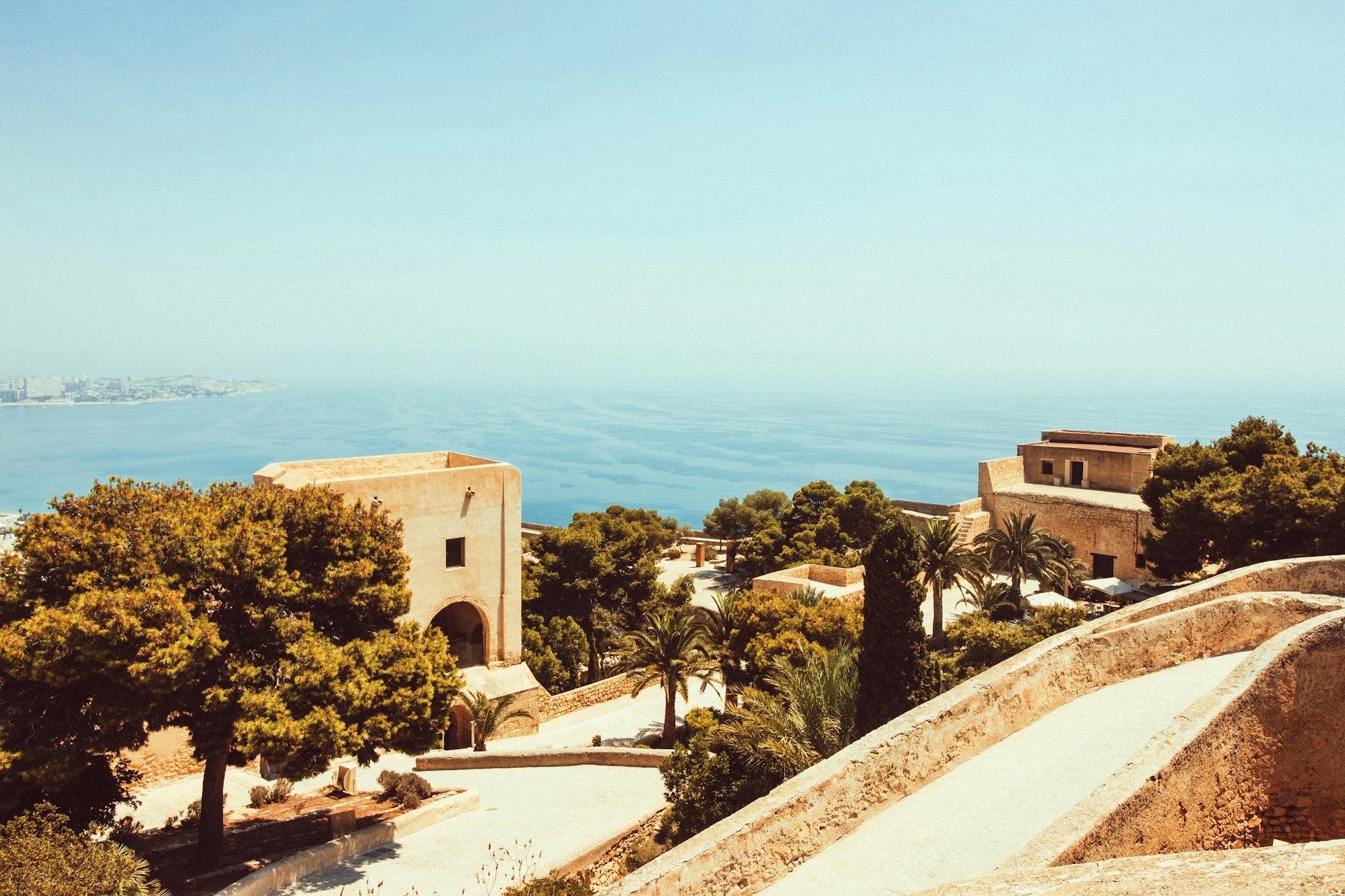 elvis-bekmanis-cOycnq2-Htc-unsplash View of the Mediterranean coastline and Málaga city from a sun-lit hilltop, framed by pine and palm trees and old stone ramparts