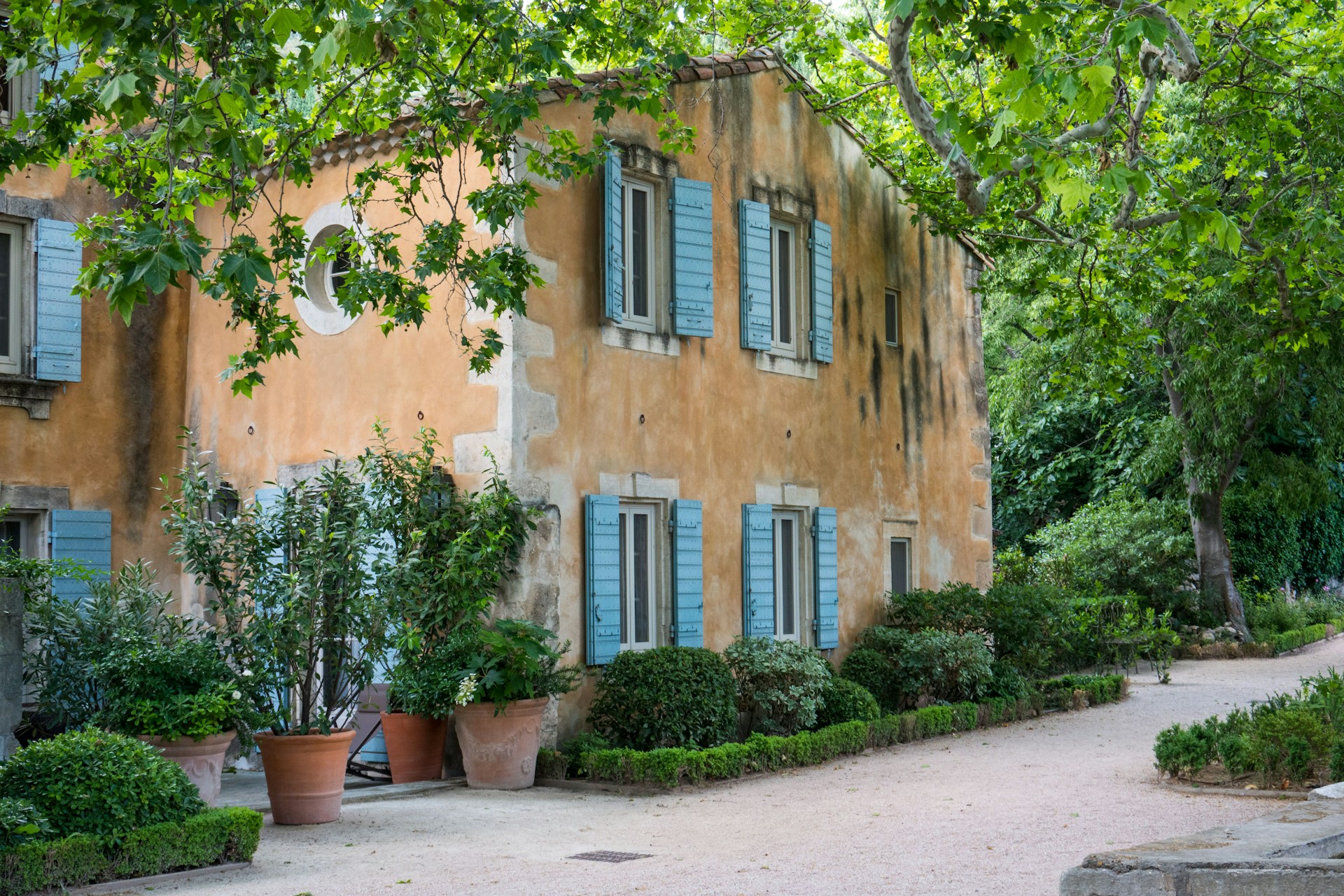 jean-nicolas_Provence @unsplash Ochre-rendered Provençal farmhouse with pale blue shutters, plane trees and terracotta pots beside a gravel courtyard