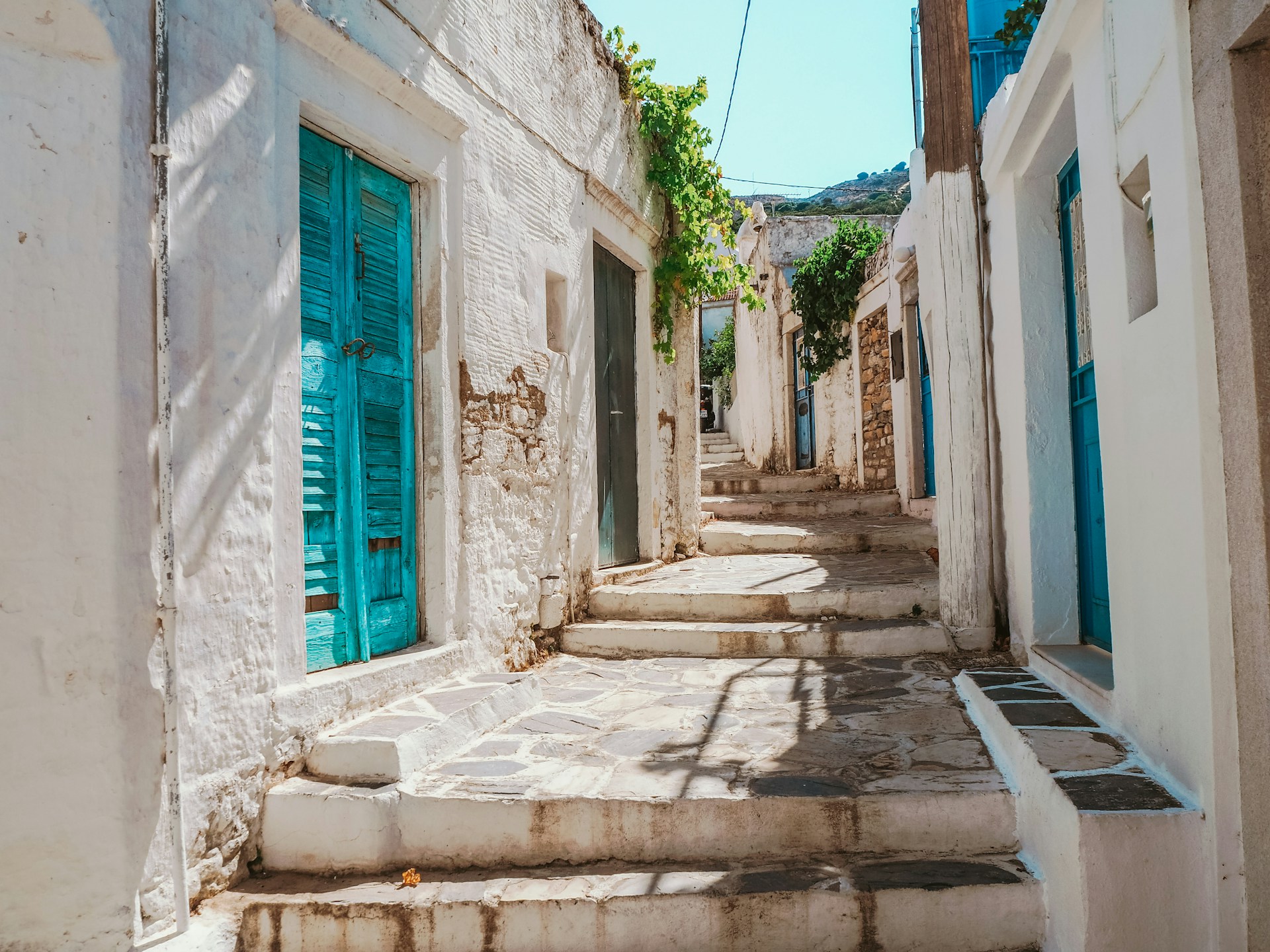 johnny-africa-Naxos@unsplash Whitewashed Cycladic village lane with turquoise doors, flagged steps and a vine trained overhead in bright sun