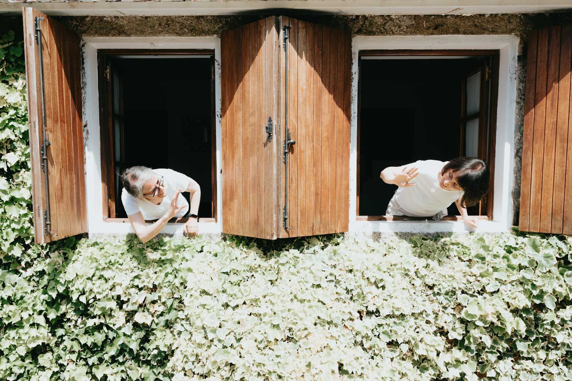 a-c-pjc-r0lSarM-unsplash Two people leaning out of neighbouring windows to chat, on an ivy-covered building with wooden shutters