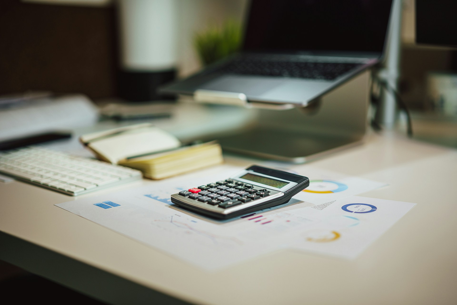 jakub-zerdzicki-8wLZi9OhsWU-unsplash Calculator resting on a printed chart on a desk, with a laptop, keyboard and notebook behind it